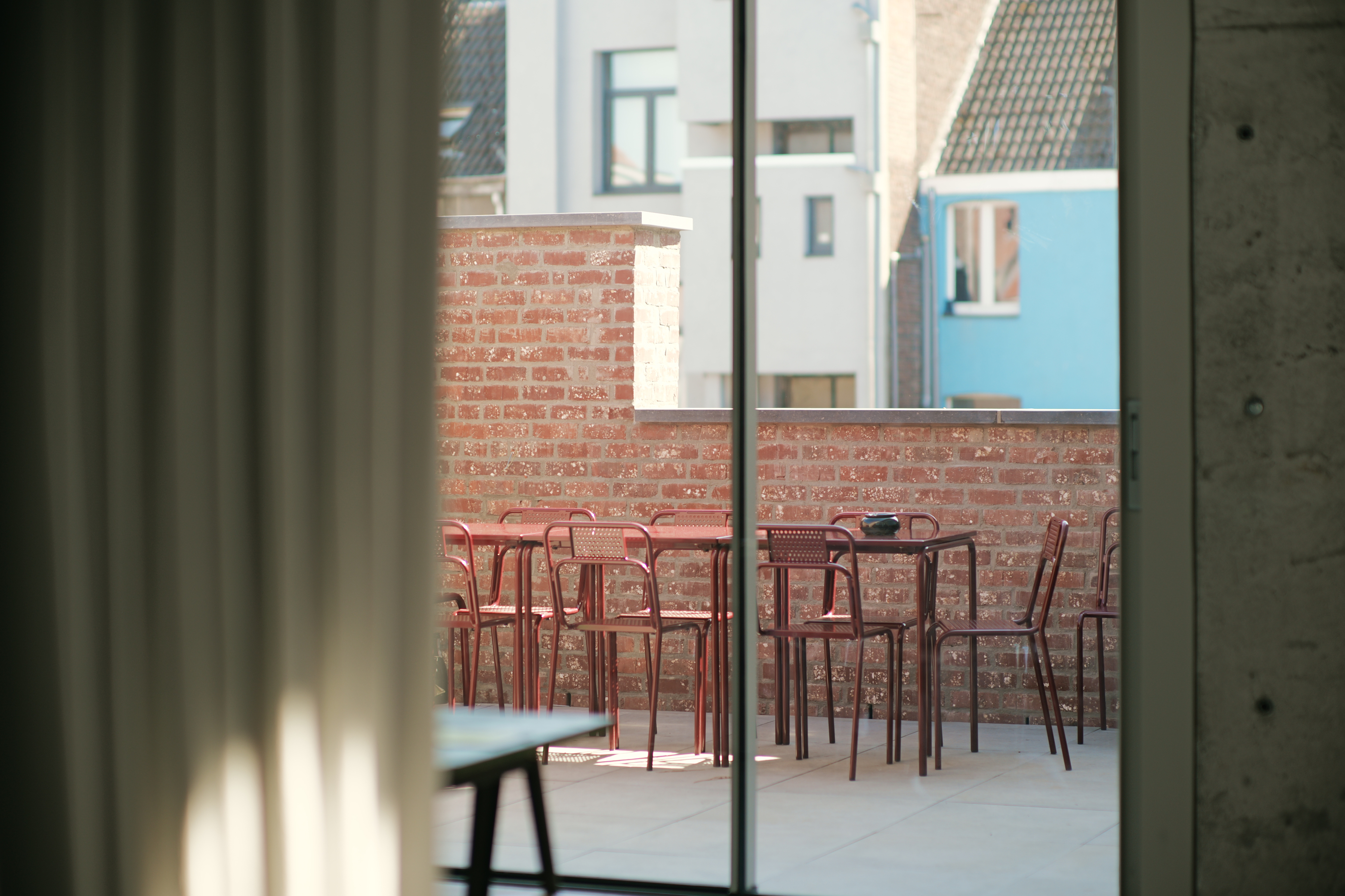 Outdoor terrace with red metal tables and chairs, surrounded by a brick wall, viewed through a glass door.