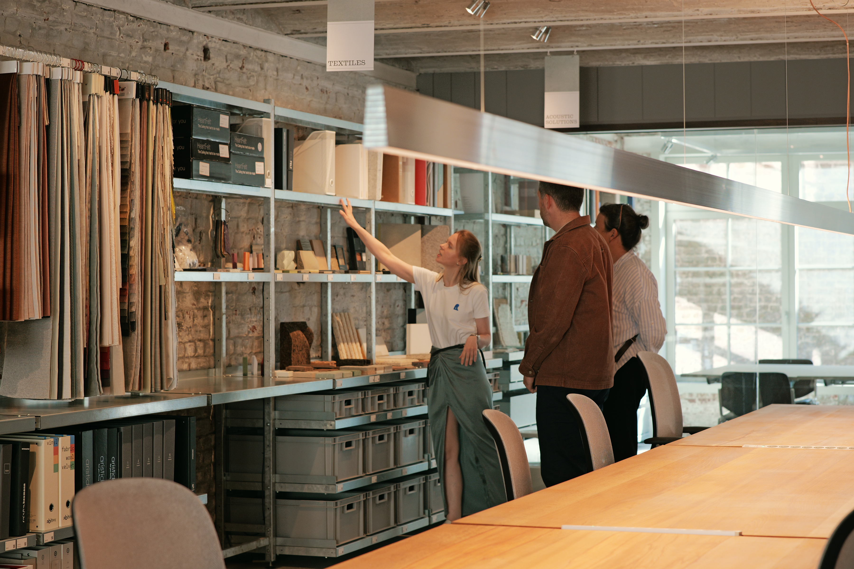Person pointing at shelves with textile and material samples while explaining to two others in a design studio library.