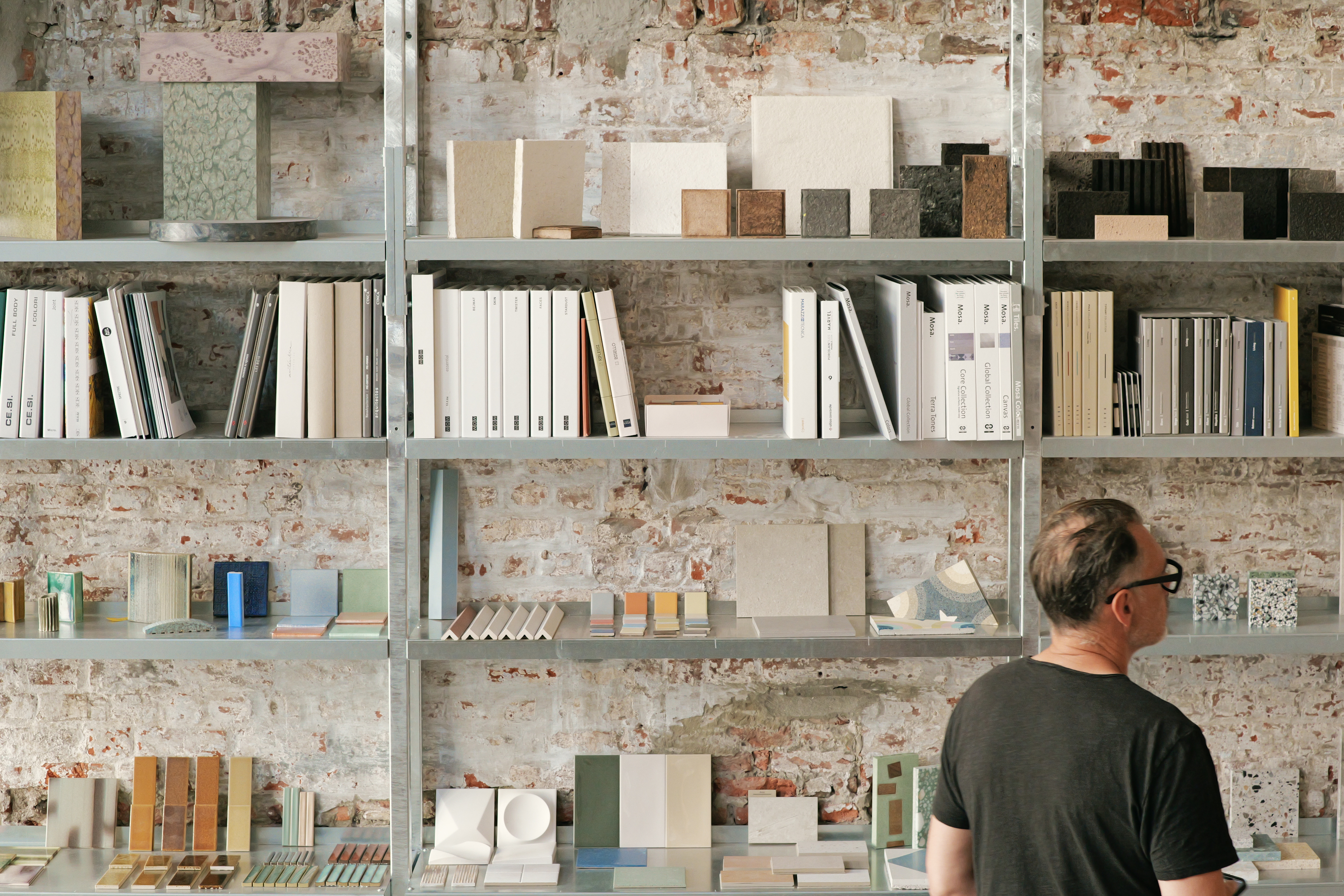 Person looking at shelves filled with material samples, books, and binders in a design studio with exposed brick walls.