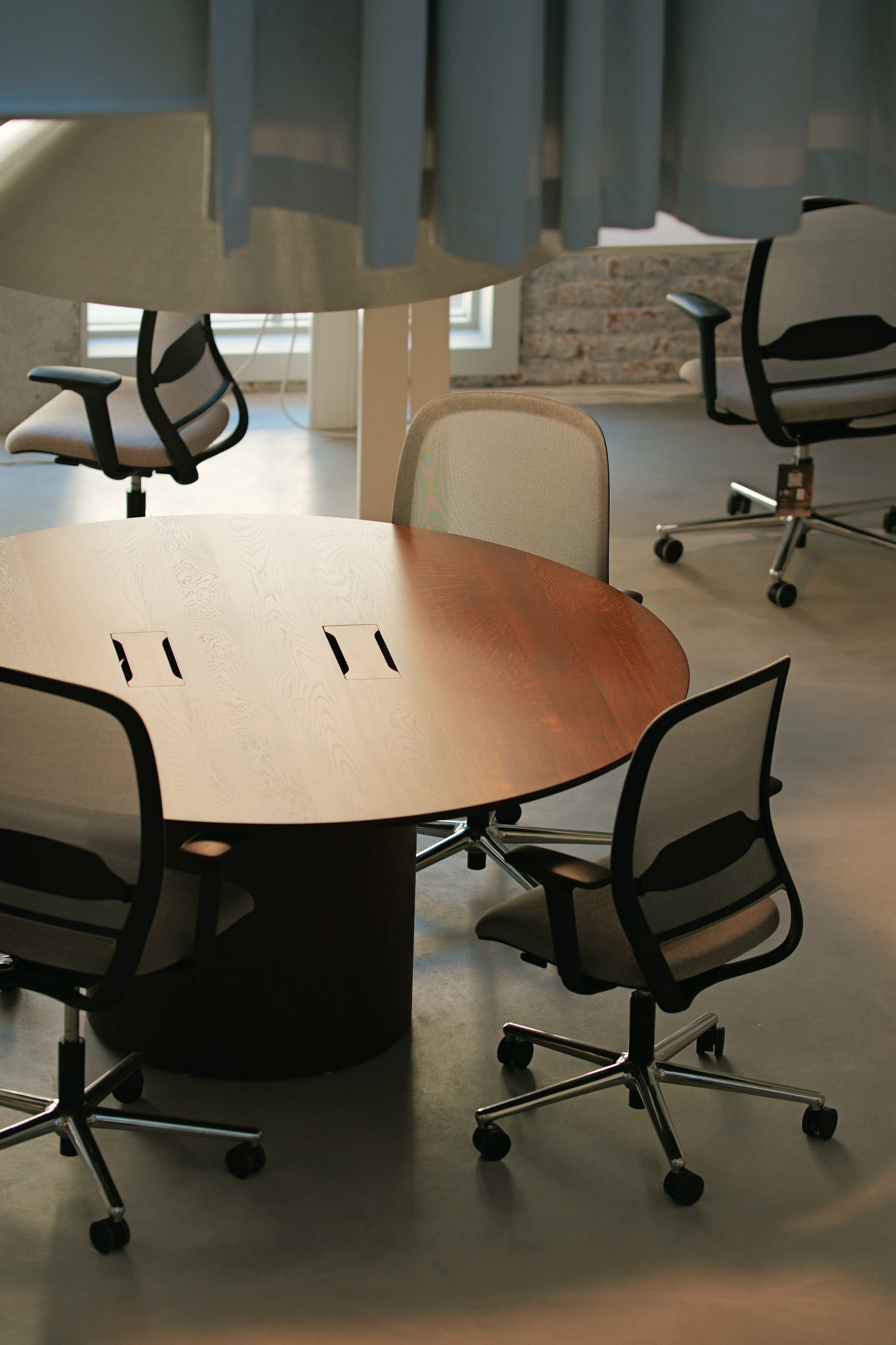 Round wooden meeting table surrounded by office chairs on wheels in a modern workspace with natural light.