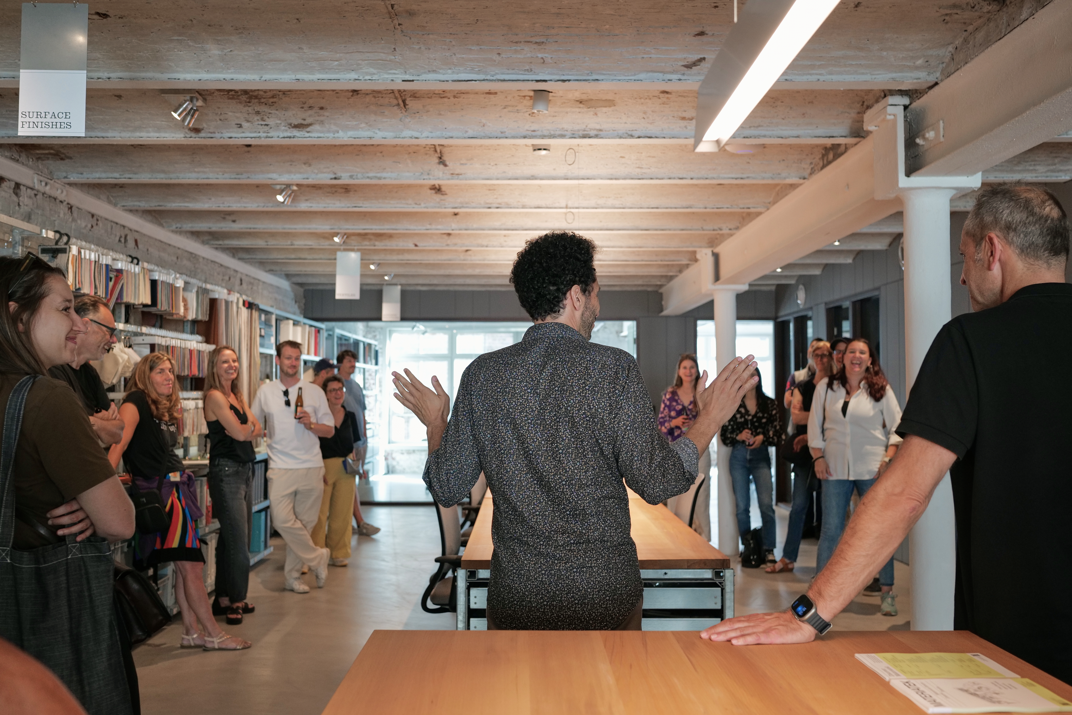 A man speaks to a group of people in a modern interior design studio, standing in front of shelves filled with surface finish samples, while the audience listens and smiles.