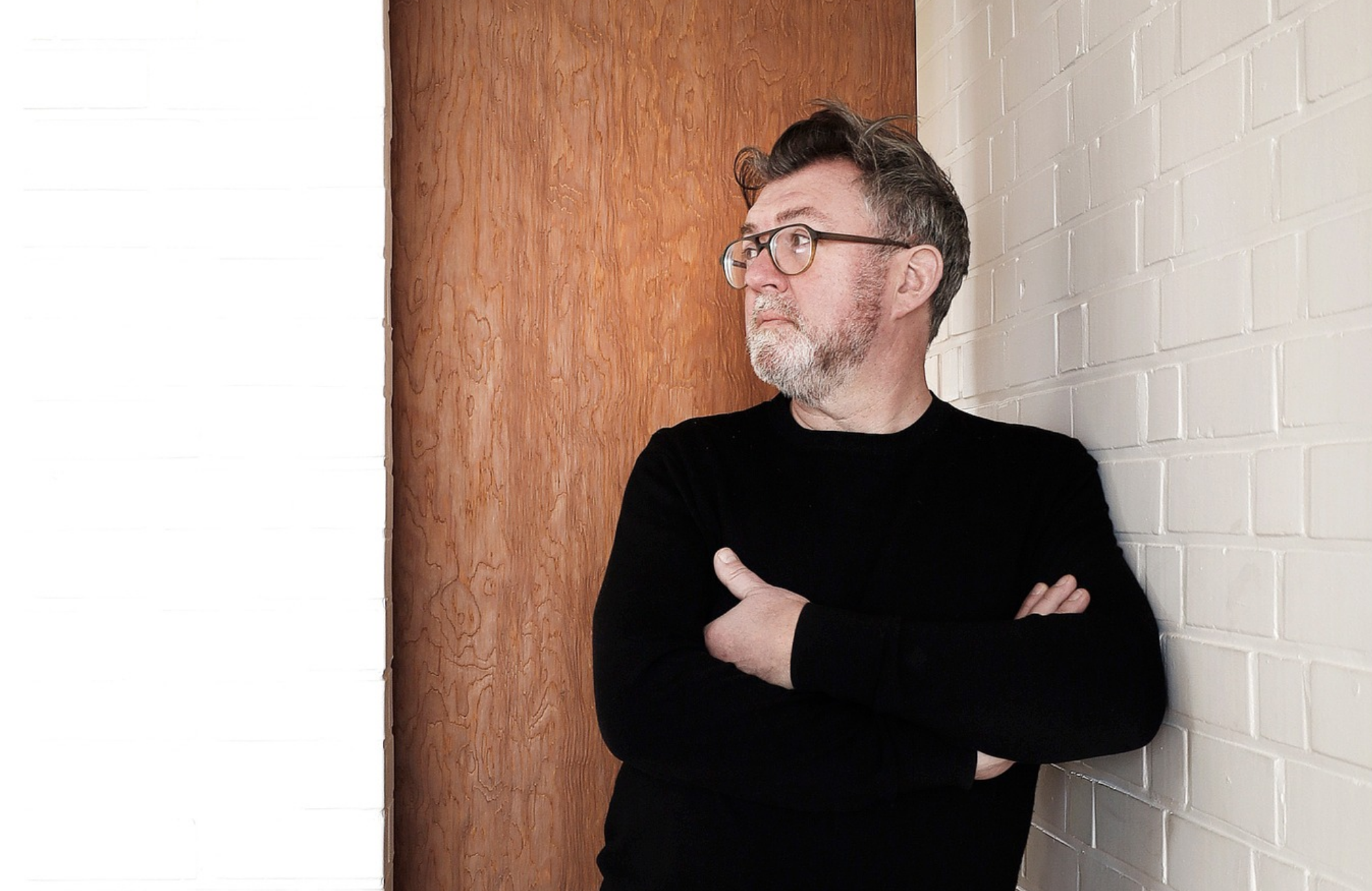 Person wearing glasses and a black sweater standing against a white brick wall and wooden panel, looking to the side.