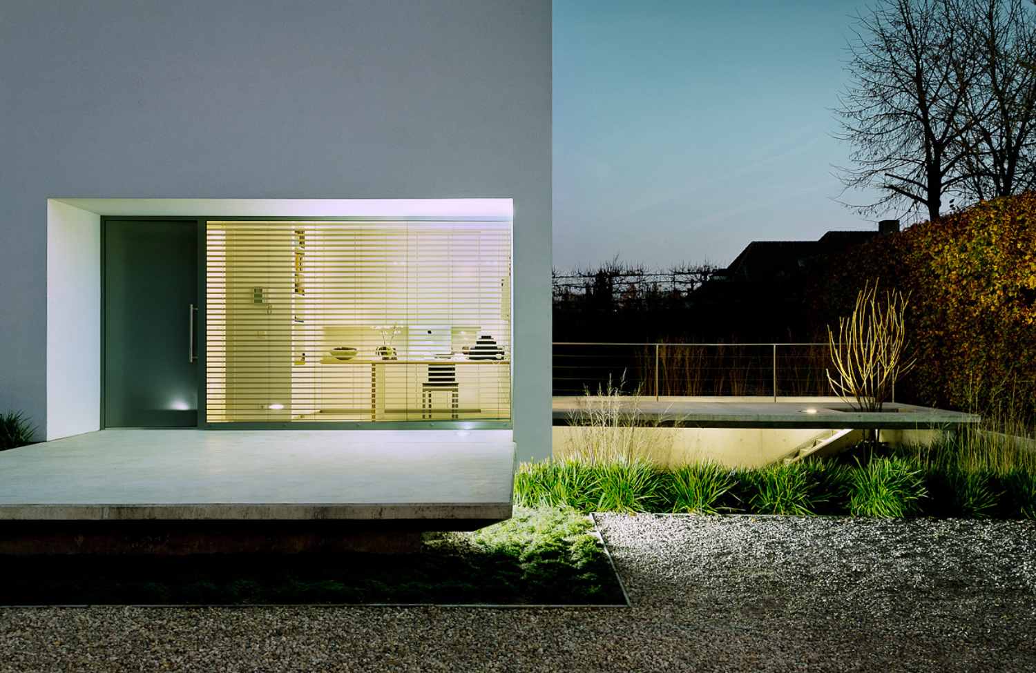Contemporary residential architecture featuring minimalist design, large glazed façade with blinds, concrete platform, and landscaped garden lighting at twilight.