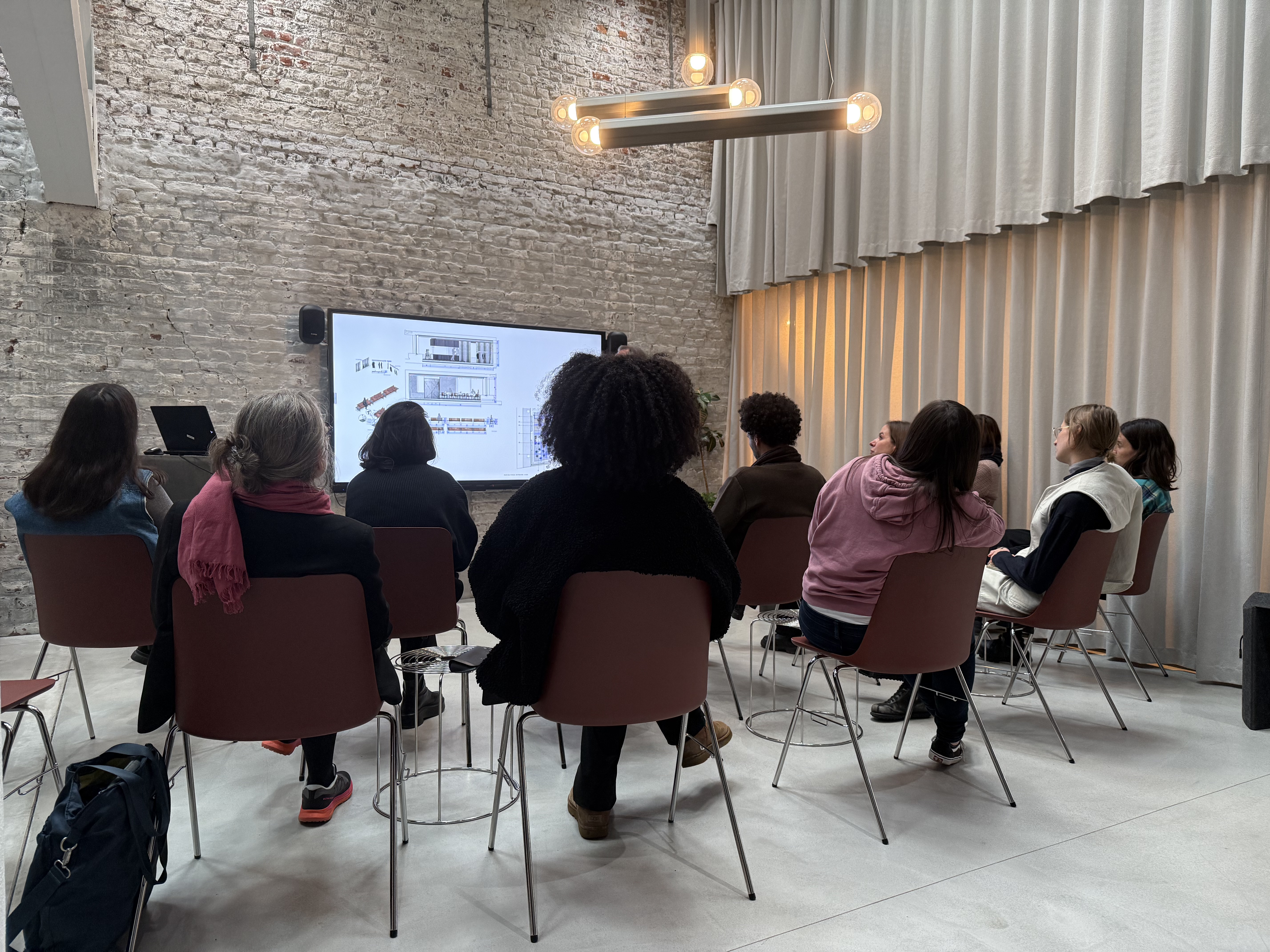 Group of people seated in a minimalist event space, watching a presentation on a large wall-mounted screen against exposed brick walls and soft grey curtains