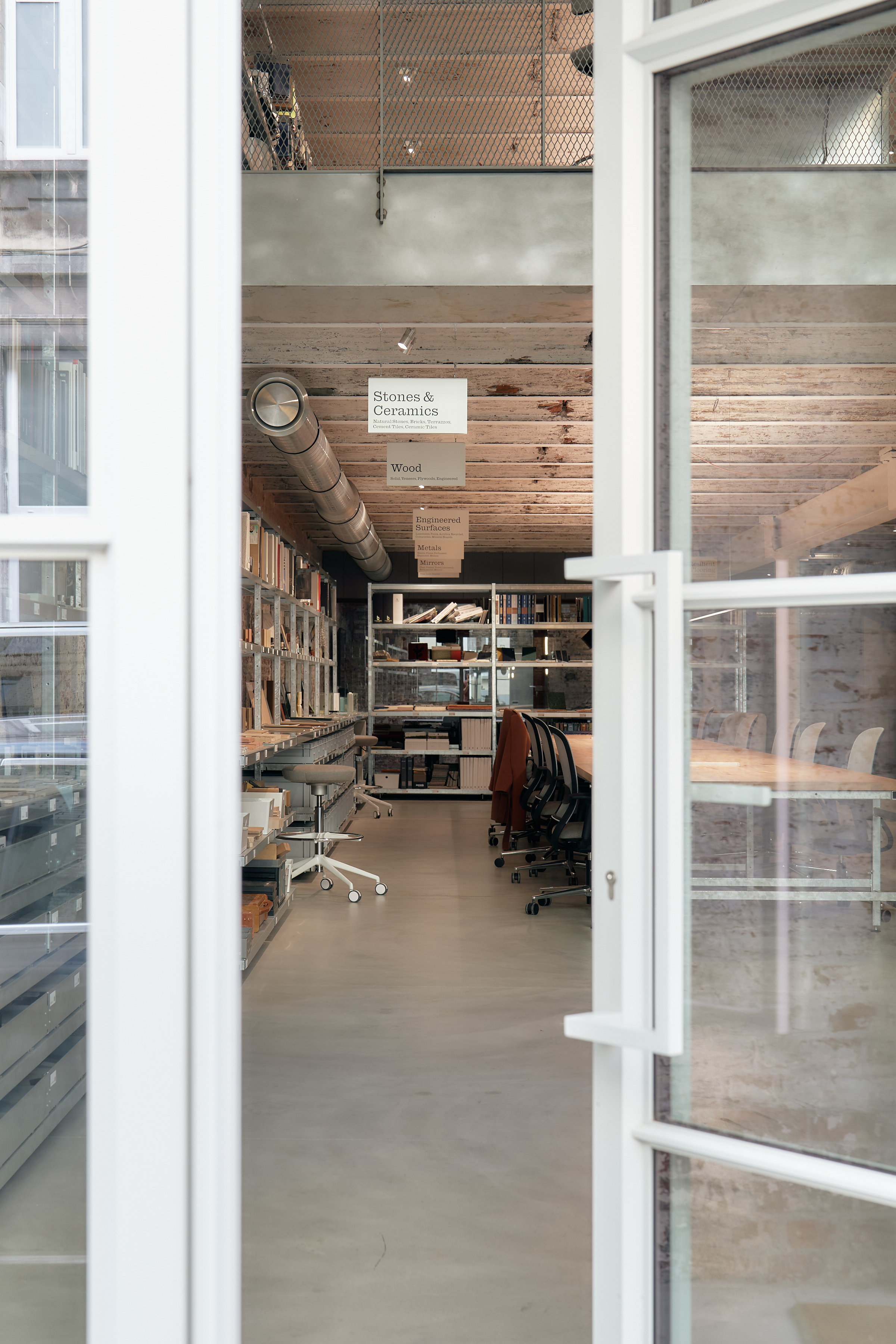 iew through glass doors into a material library with labeled sections for stones, ceramics, wood, and engineered surfaces, featuring long worktables, shelving, and industrial-style interiors.