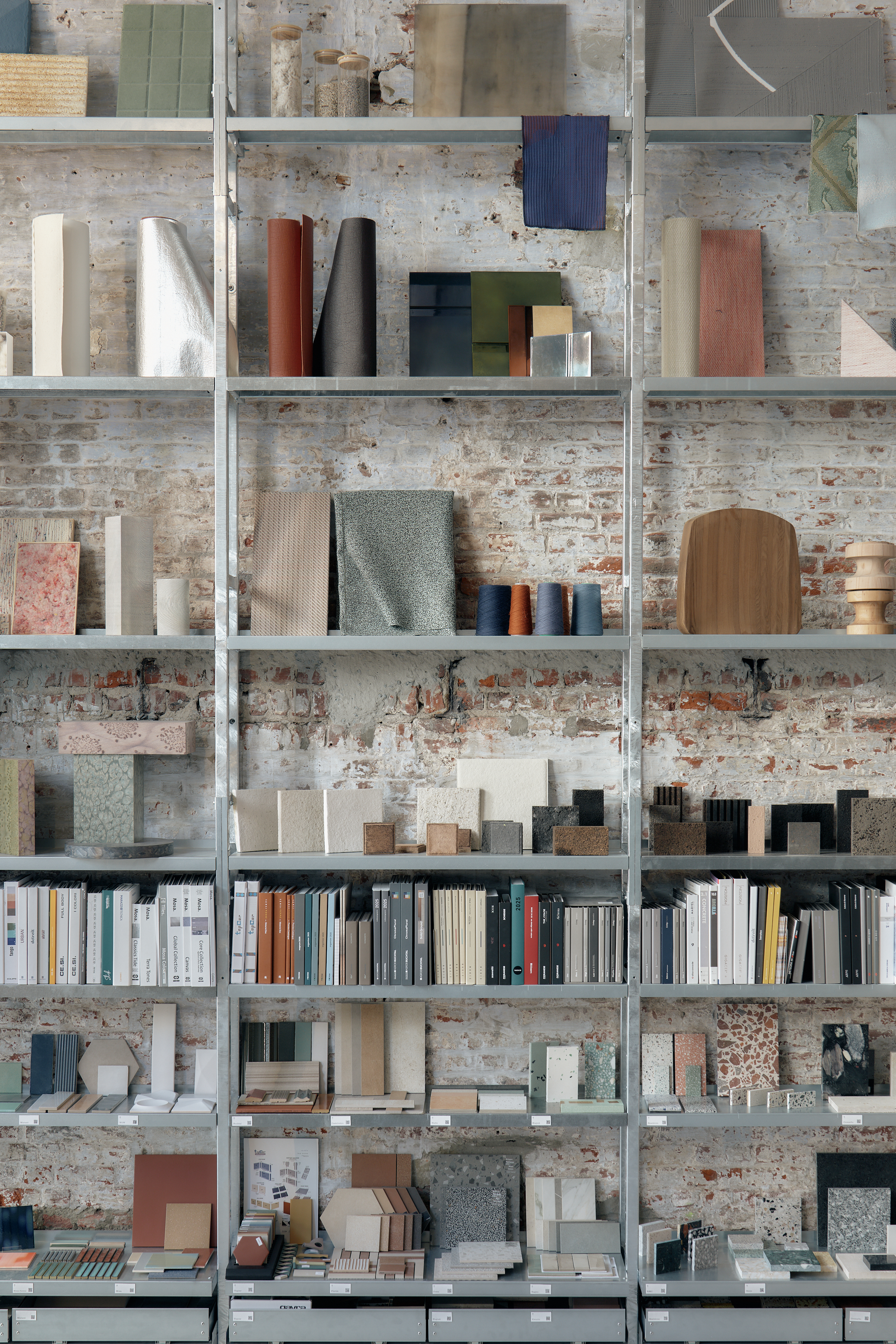 Shelves displaying a curated collection of architectural materials, textures, books, and samples against a raw brick wall.